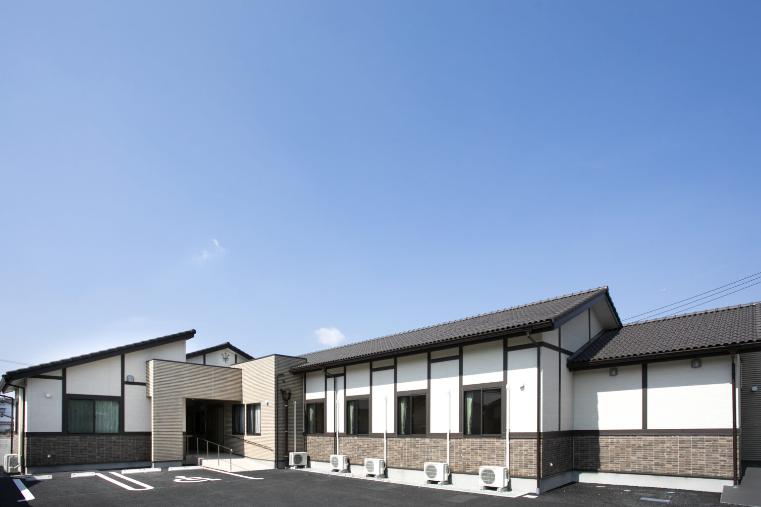 介護施設の建物外観。モダンな平屋造りで、白と茶色の外壁、広い駐車スペースがある。